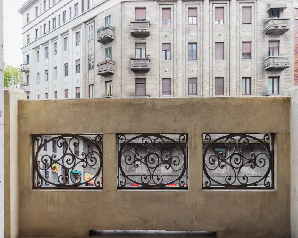 Pequeño balcón urbano con barandilla de hierro forjado decorativa y vista a las fachadas de los edificios cercanos.
