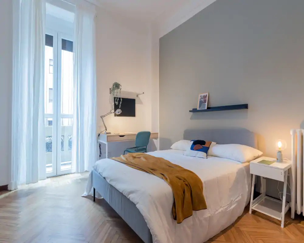 Dormitorio luminoso y con estilo con una cama doble cómoda, mesita de noche y escritorio junto a la alta ventana que aporta abundante luz natural.