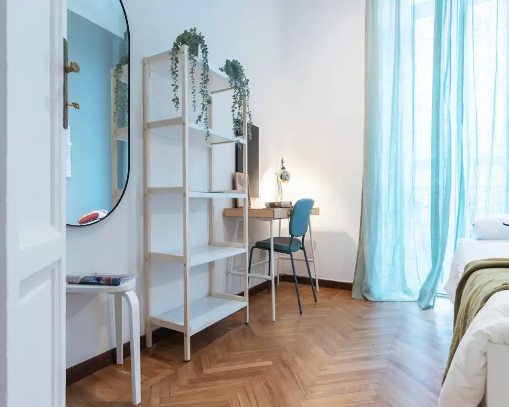 Angolo camera luminoso e minimal con bordo del letto visibile, una scrivania in legno e scaffalatura. La luce naturale filtra attraverso tende azzurro pallido creando un'area lavoro tranquilla accanto al letto.