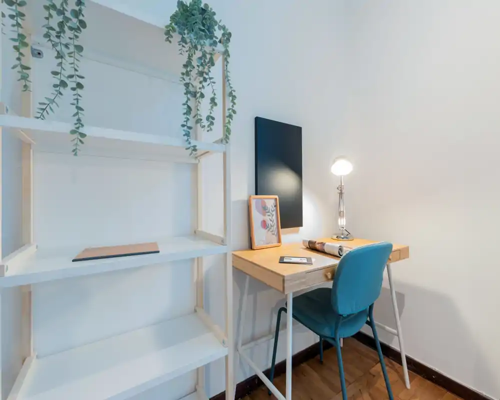 Angolo studio luminoso e minimal con scrivania in legno, sedia turchese, lampada da tavolo e piante decorative che creano un ambiente di lavoro tranquillo.