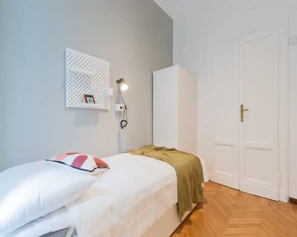 Camera singola compatta e luminosa con letto ben rifatto, tessili morbidi e un armadietto minimal. Toni neutri e pavimento in legno caldo creano un'atmosfera tranquilla ideale per annunci di affitto.