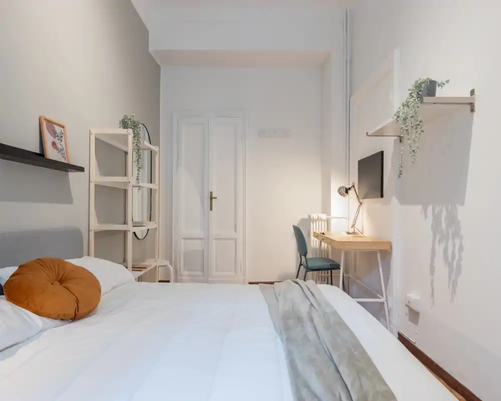 Dormitorio luminoso y moderno con una cama grande en primer plano, textiles suaves, un escritorio con lámpara y estanterías minimalistas: un espacio tranquilo y acogedor ideal para anuncios.