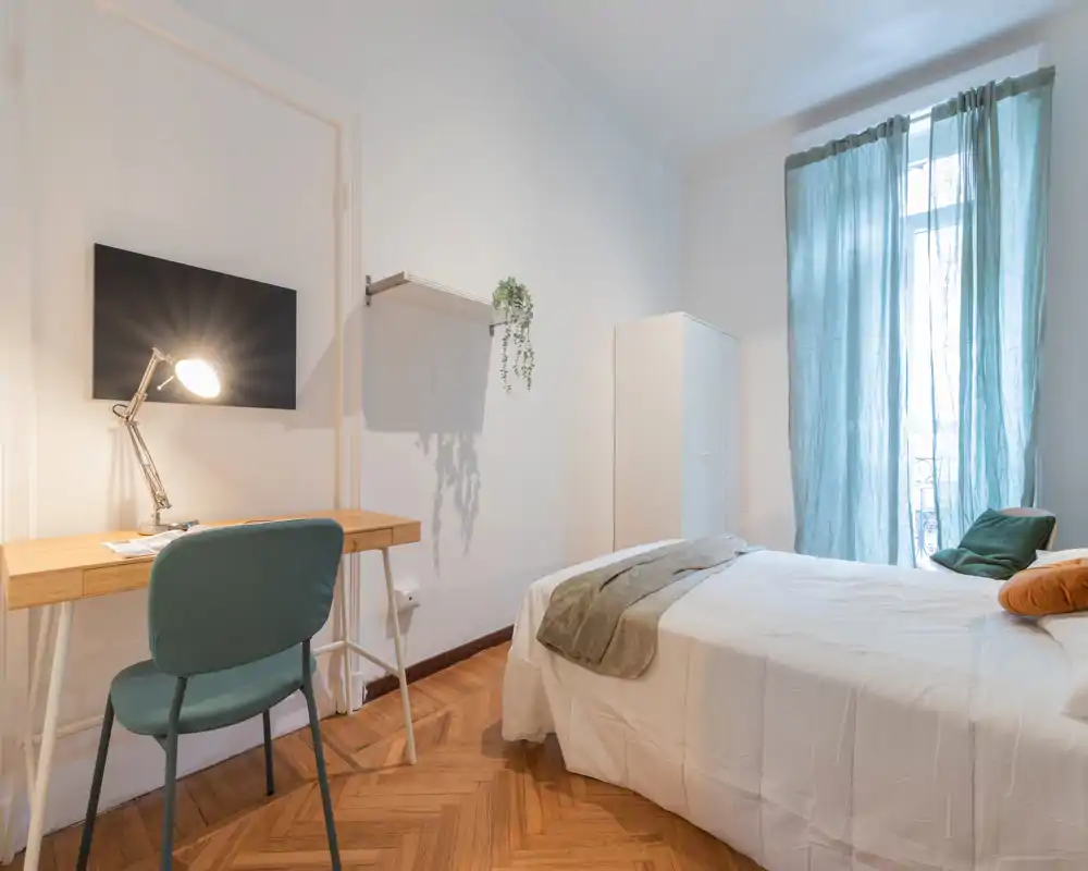 Dormitorio moderno y luminoso con cama doble cómoda, ropa de cama suave y un escritorio ordenado junto a la pared; gran ventana con cortinas que aporta luz natural.