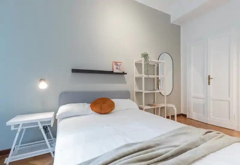 Dormitorio moderno y luminoso con cama doble bien hecha, mesita minimalista y estantería; tonos neutros y un cojín redondo crean un ambiente acogedor.