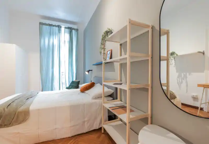 Dormitorio luminoso y bien ambientado con cama completa, ropa de cama suave y cojines decorativos; una estantería y un gran espejo aportan profundidad y almacenamiento.