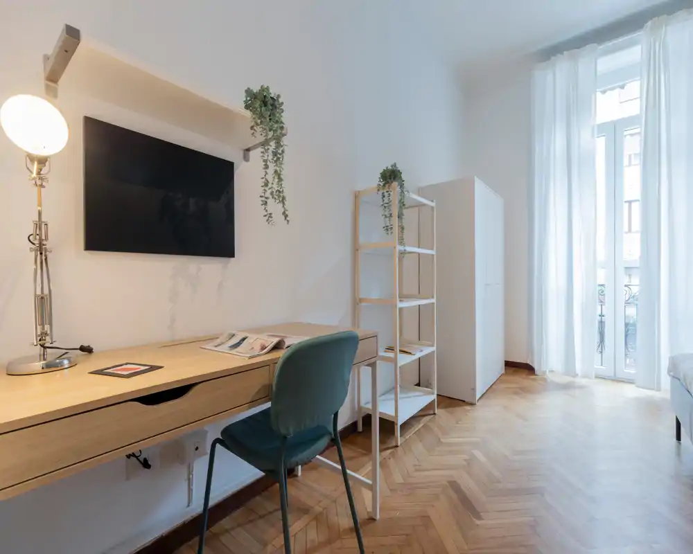 Dormitorio luminoso y minimalista con escritorio de madera, silla verde y una gran ventana que aporta luz natural; estantería y armario sencillos sobre suelo de parquet claro.