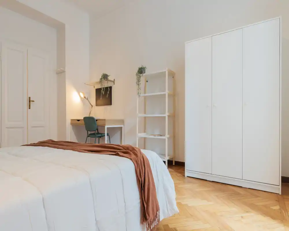 Dormitorio luminoso y bien presentado con cama grande en primer plano, armario blanco y un pequeño escritorio con estantería — ideal para anuncios que destaquen confort y espacio de trabajo.