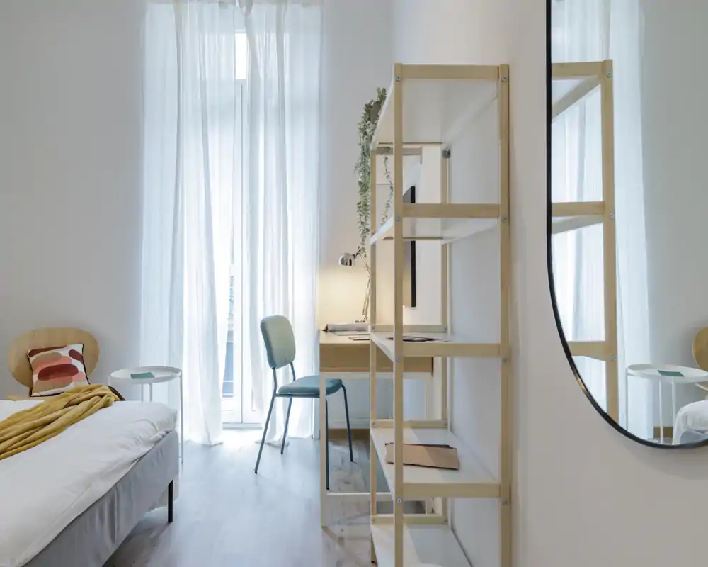 Dormitorio luminoso y minimalista con cama bien hecha, cortinas traslúcidas que iluminan la habitación, escritorio pequeño y estanterías — ideal para una estancia tranquila o para trabajar.