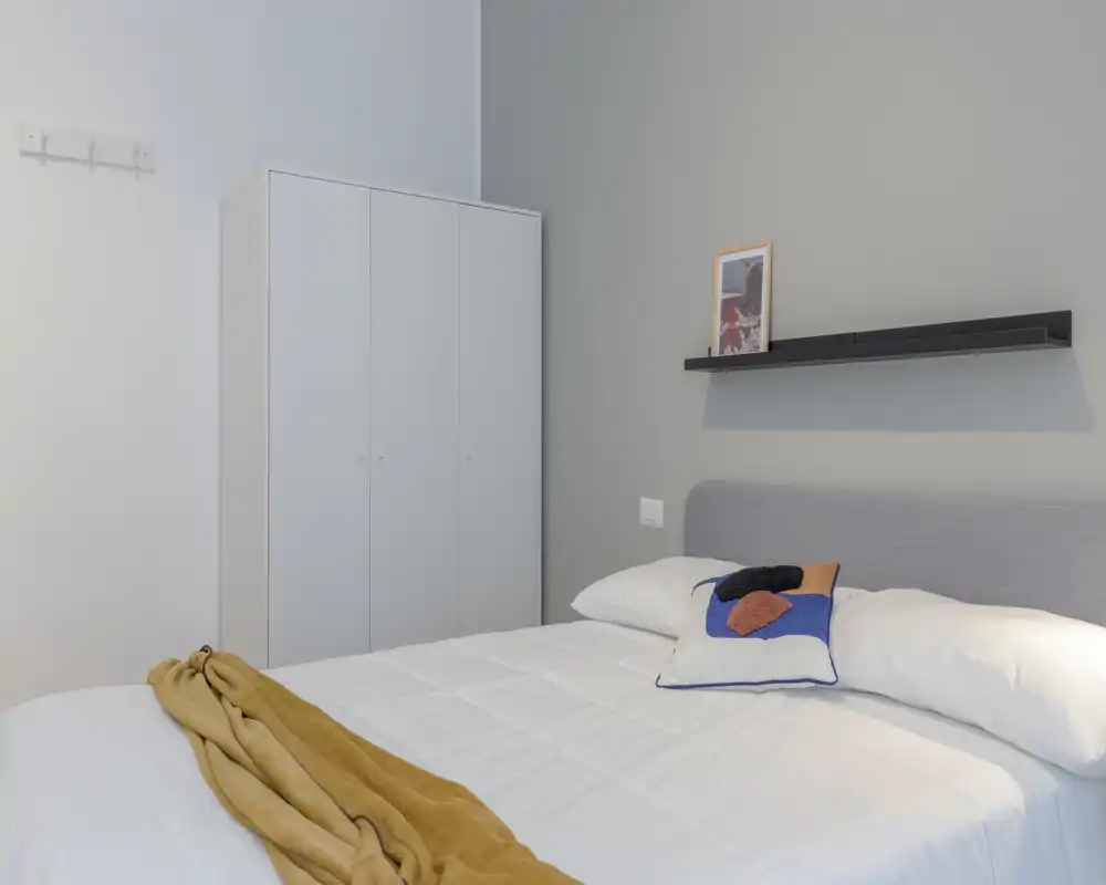 Dormitorio moderno y luminoso con cama doble bien hecha, cojín decorativo y armario. Tonos neutros y una repisa sencilla ofrecen un ambiente tranquilo y acogedor perfecto para un anuncio.