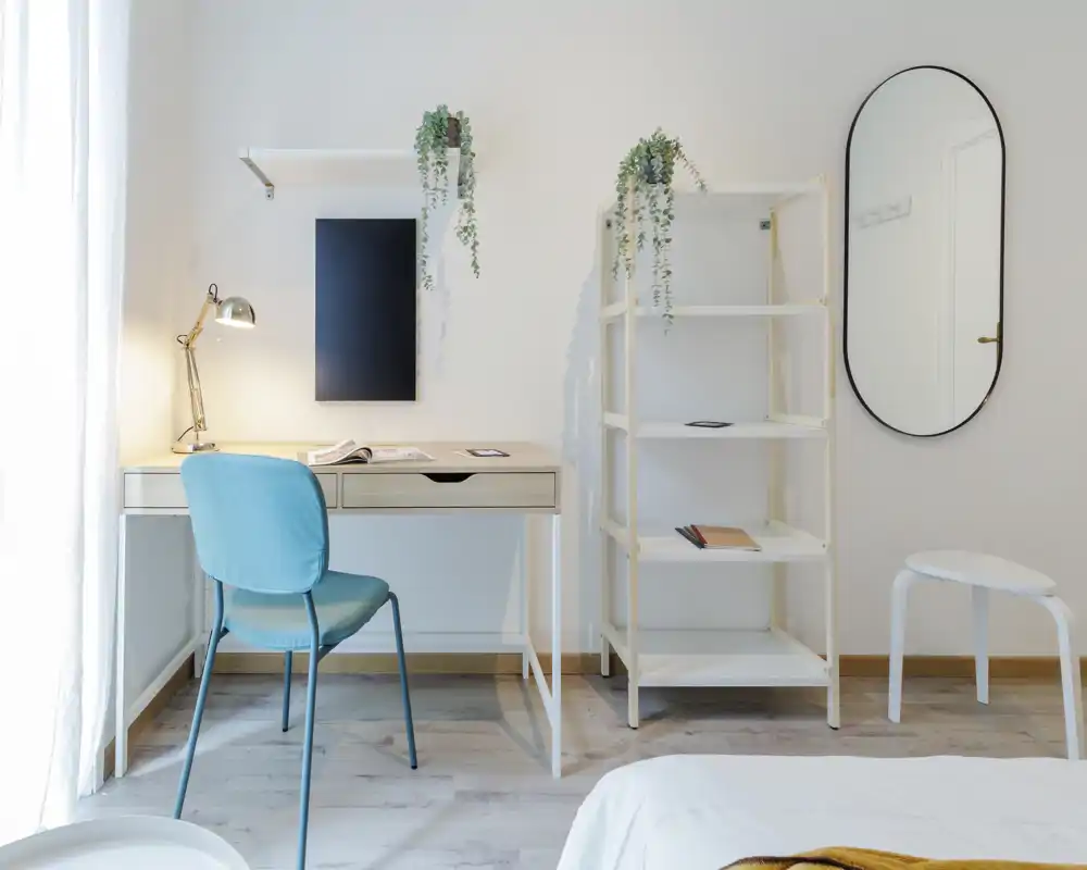 Rincón de dormitorio luminoso y minimalista con escritorio, silla y vista parcial de la cama. Luz natural, plantas y detalles suaves crean un ambiente tranquilo y funcional para trabajar y descansar.