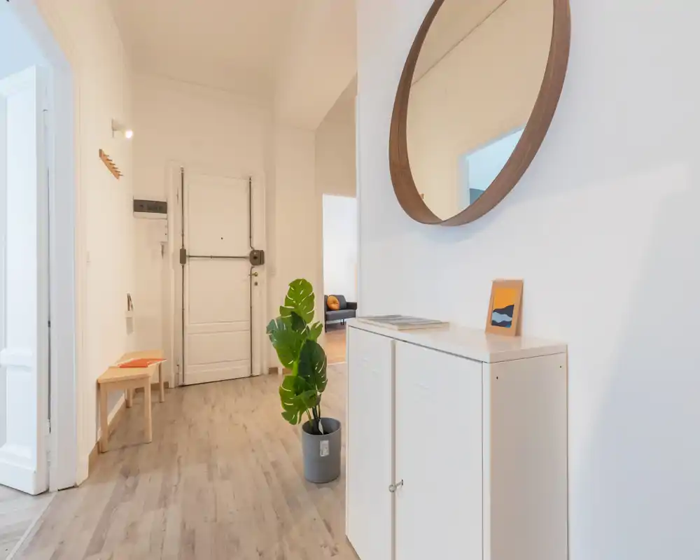 Entrada luminosa y minimalista con espejo redondo decorativo, un mueble pequeño y un banco — estilo moderno y suelos efecto madera.