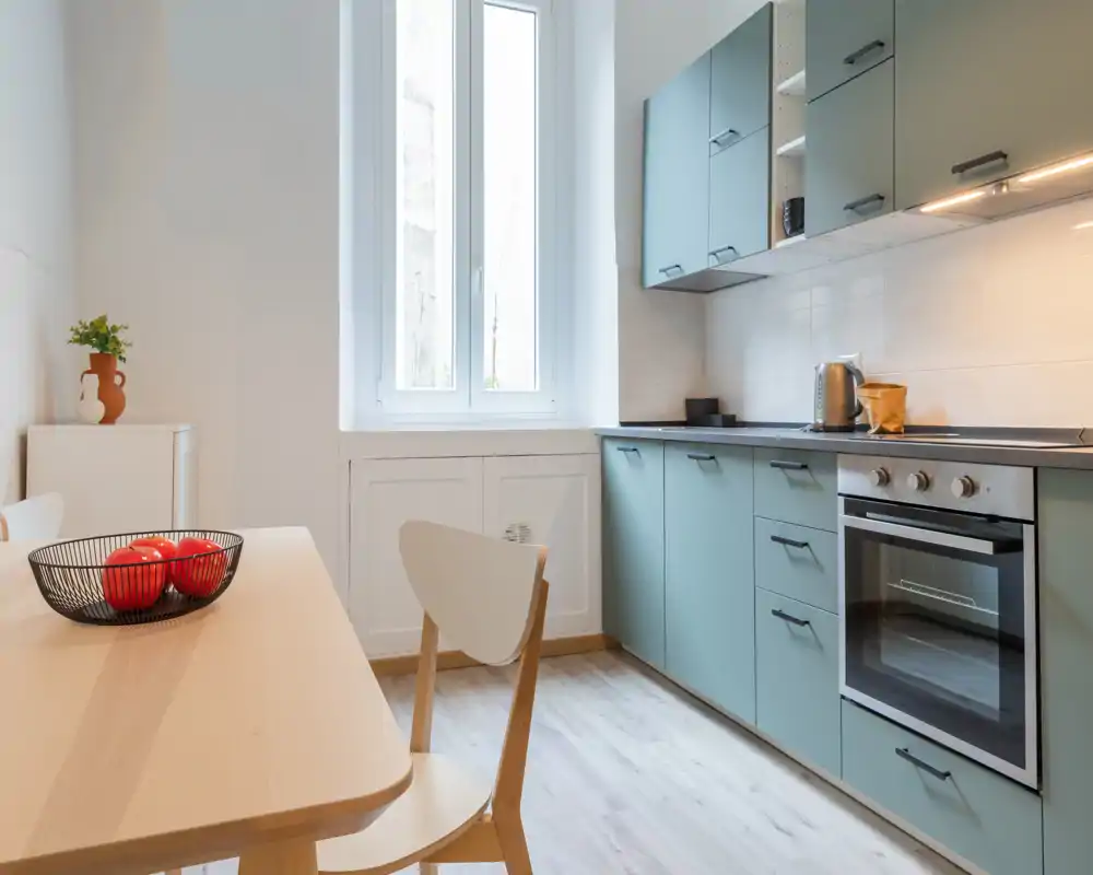 Cocina moderna y luminosa con armarios verde suave, horno integrado y mesa de madera clara. La luz natural inunda el espacio, creando un área de cocina limpia y acogedora.