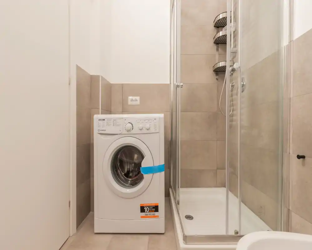 Baño compacto con lavadora de carga frontal junto a una ducha con mampara de vidrio; azulejos beige neutro y distribución limpia y funcional.