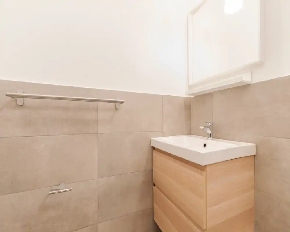 Esquina de baño moderno con mueble de lavabo suspendido, grifo cromado, paredes alicatadas y espejo encima; luminoso, limpio y minimalista.