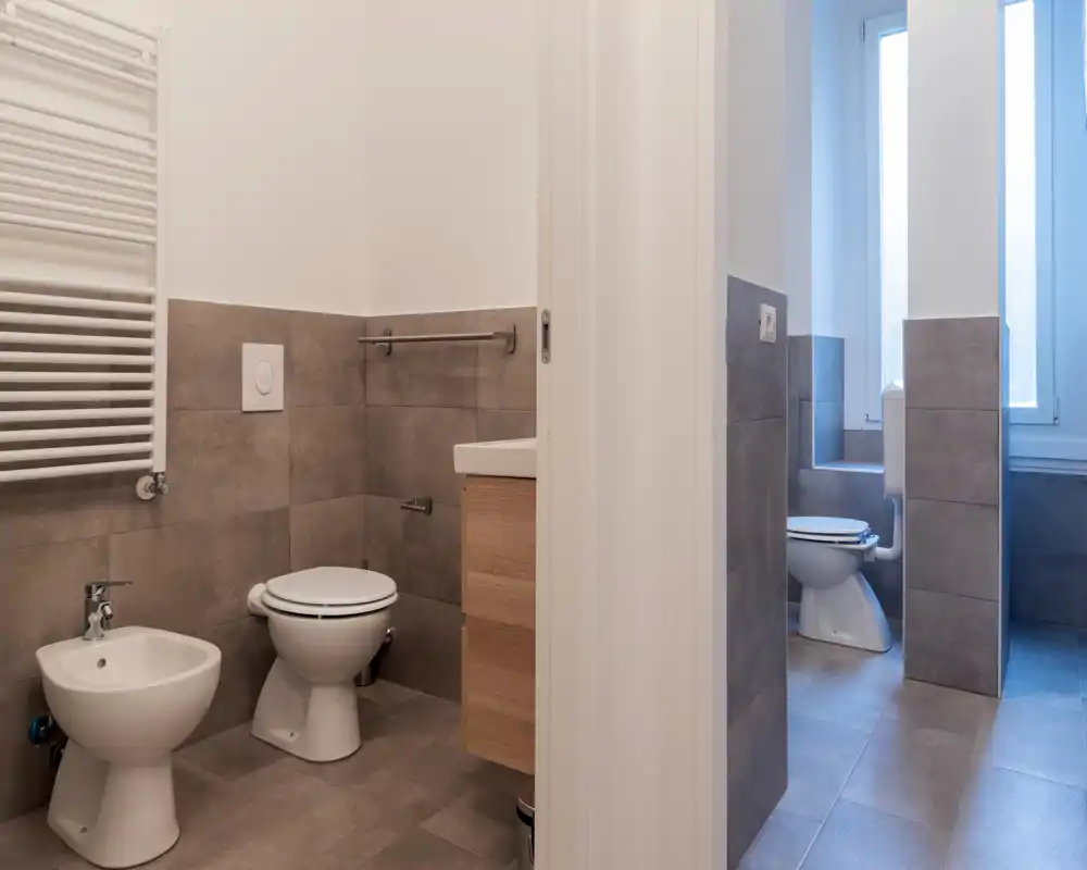 Baño moderno y bien iluminado con inodoro, bidé y mueble de lavabo; azulejos grises neutras y una ventana alta que aporta luz natural.