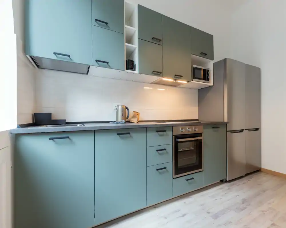 Cocina moderna y luminosa con armarios verde menta, horno integrado, microondas y frigorífico de acero. Líneas limpias y luz bajo los muebles que crean un área de cocina ordenada y funcional.