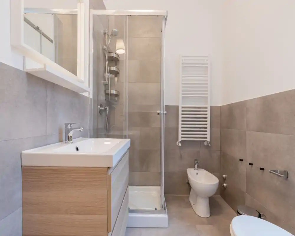 Baño moderno y bien iluminado con ducha con mampara de cristal, radiador toallero mural, lavabo con mueble, bidé y inodoro en tonos neutros de azulejos.