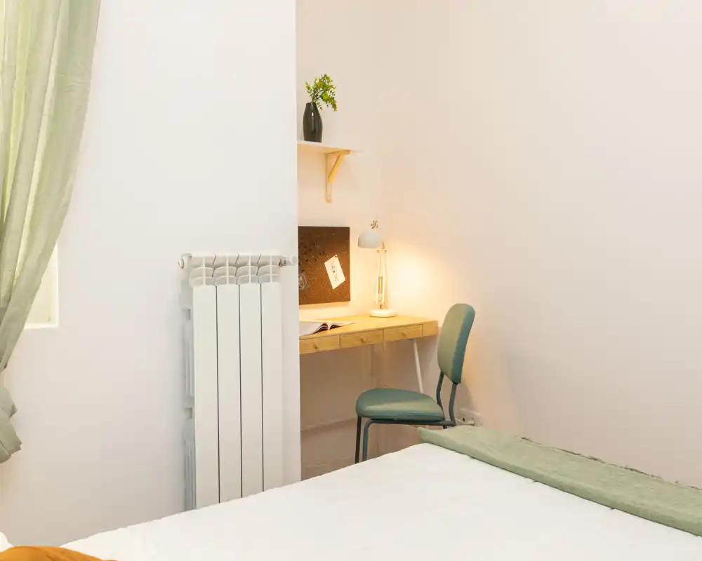Angolo camera luminoso e semplice con letto visibile, piccolo scrittoio in legno, sedia e lampada: toni neutri e delicati accenti verdi.