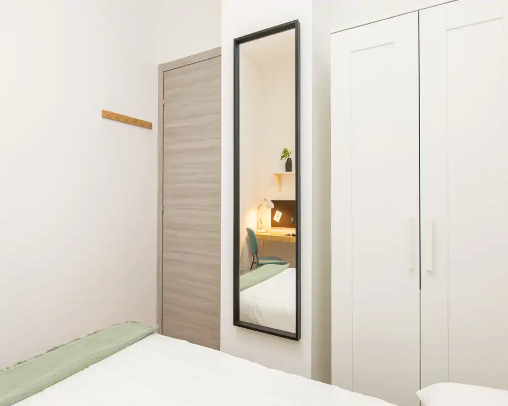 Camera da letto compatta e luminosa con letto ben rifatto, uno specchio alto e un armadio bianco; un piccolo angolo studio è visibile nello specchio.