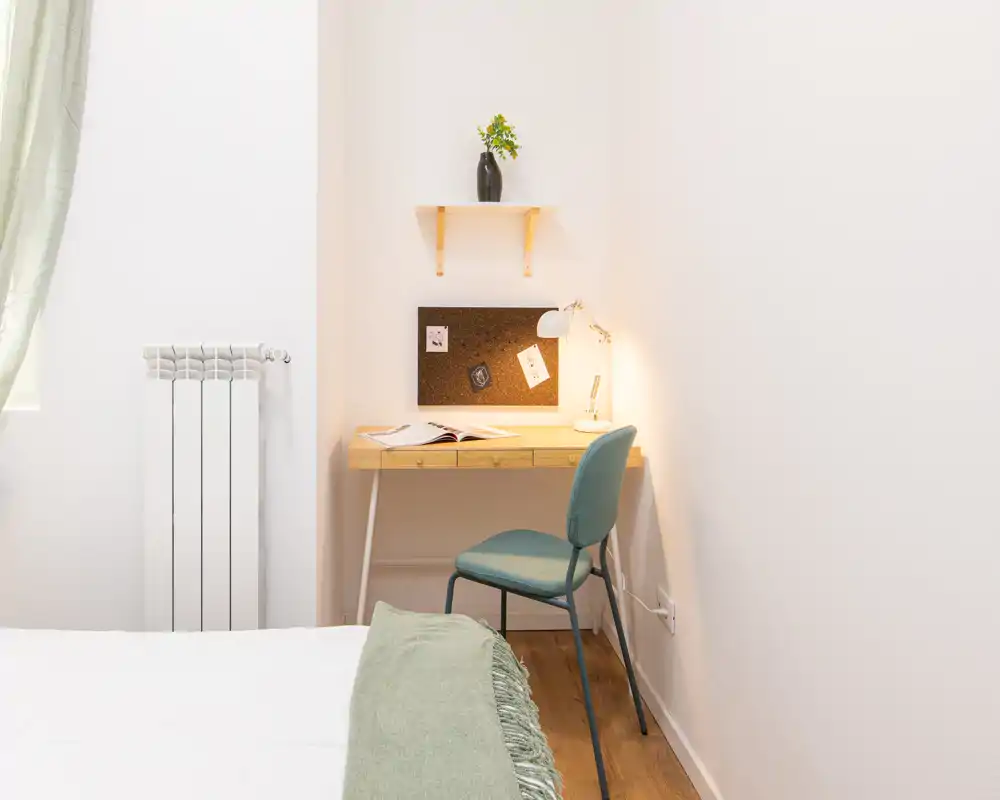 Angolo camera luminoso e minimal con bordo del letto visibile, scrivania in legno e una lampada accogliente — ideale per un’area di studio tranquilla.