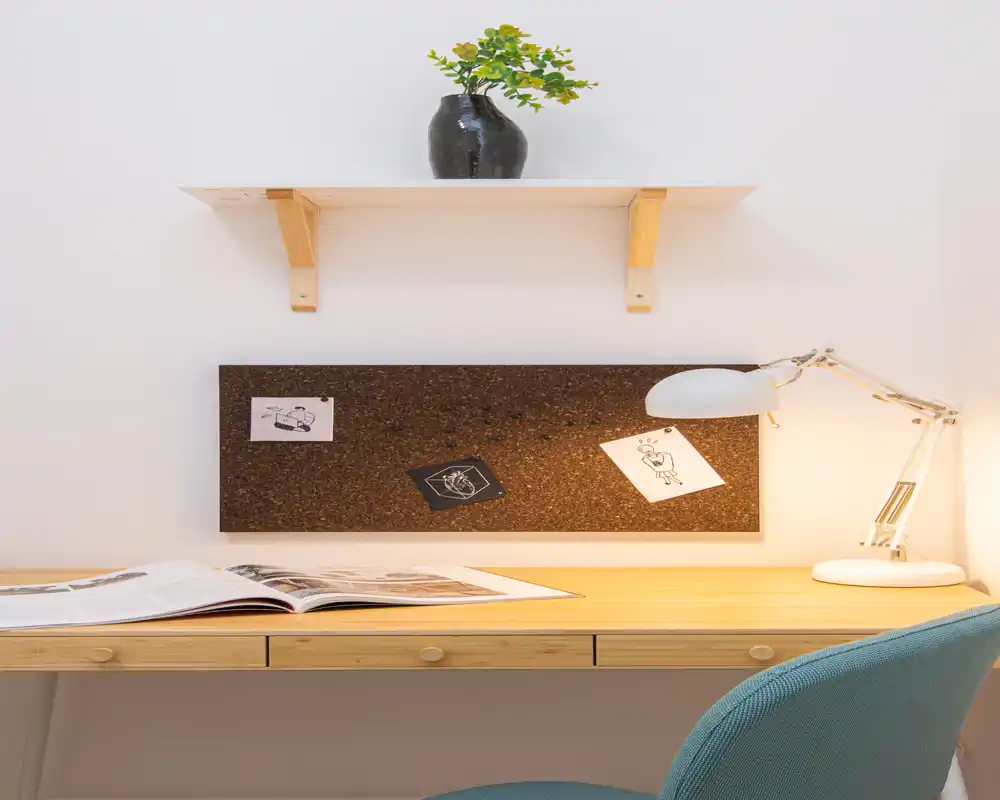 Angolo studio ordinato con scrivania in legno, lampada da tavolo, bacheca in sughero e piccolo vaso decorativo — uno spazio luminoso e tranquillo per leggere o lavorare.