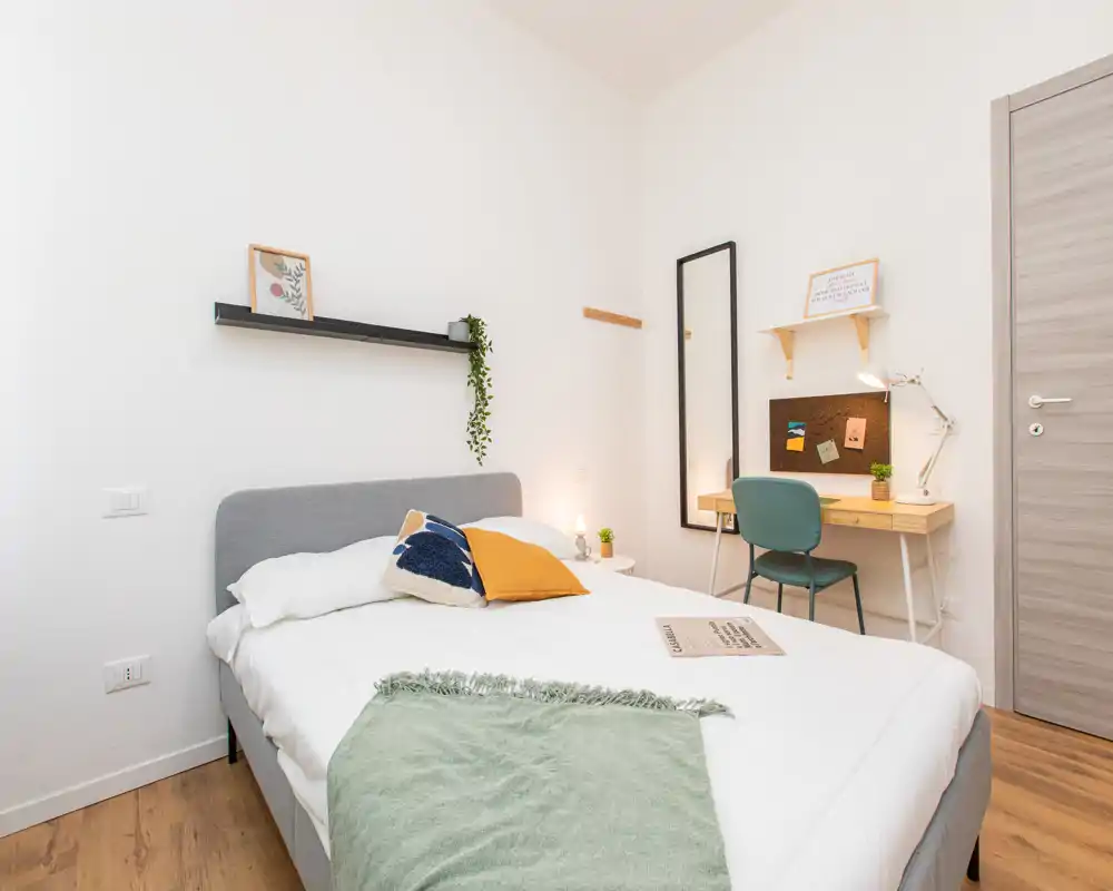 Camera da letto moderna e luminosa con letto matrimoniale, cuscini decorativi e una piccola scrivania vicino alla porta — ideale per affitti brevi o come stanza ospiti accogliente.