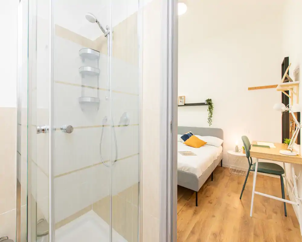 Camera da letto moderna e luminosa con letto matrimoniale visibile, comodino e una piccola postazione di lavoro. Arredo pulito, pavimento in legno caldo e buona luce naturale rendono l'ambiente accogliente.