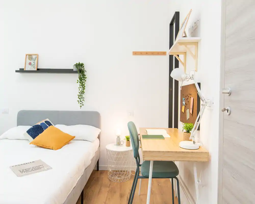 Camera da letto luminosa e moderna con comodo letto matrimoniale, cuscini decorativi e una piccola scrivania a muro — perfetta per uno studente o professionista in cerca di un ambiente accogliente e ben illuminato.