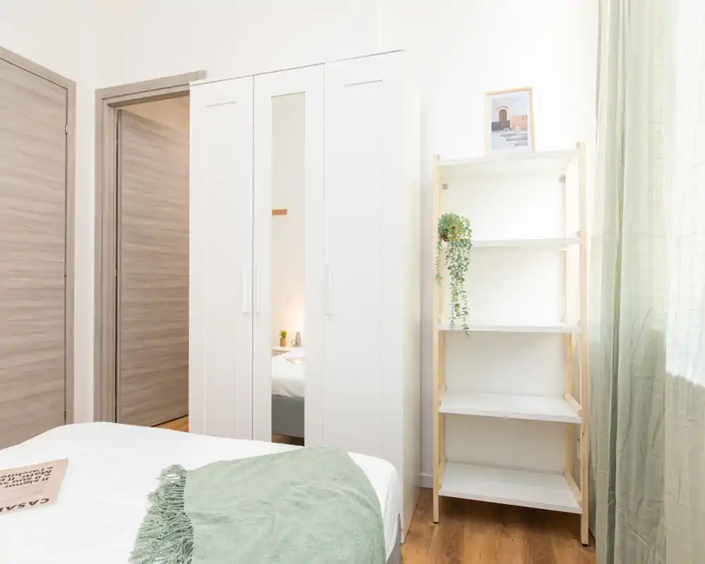 Camera da letto luminosa e minimale con un armadio bianco, una mensola aperta e un letto ben rifatto con biancheria bianca e un plaid verde. La luce naturale entra dalla finestra con tende chiare, creando un’atmosfera ariosa e rilassante perfetta per gli annunci.