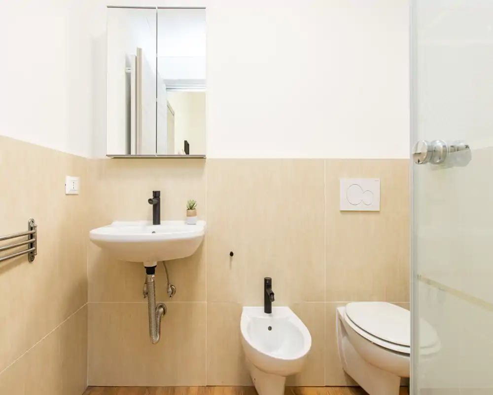 Bagno moderno e luminoso con lavabo a parete, bidet e wc, piastrelle beige chiare e mobiletto specchiato sopra il lavabo.