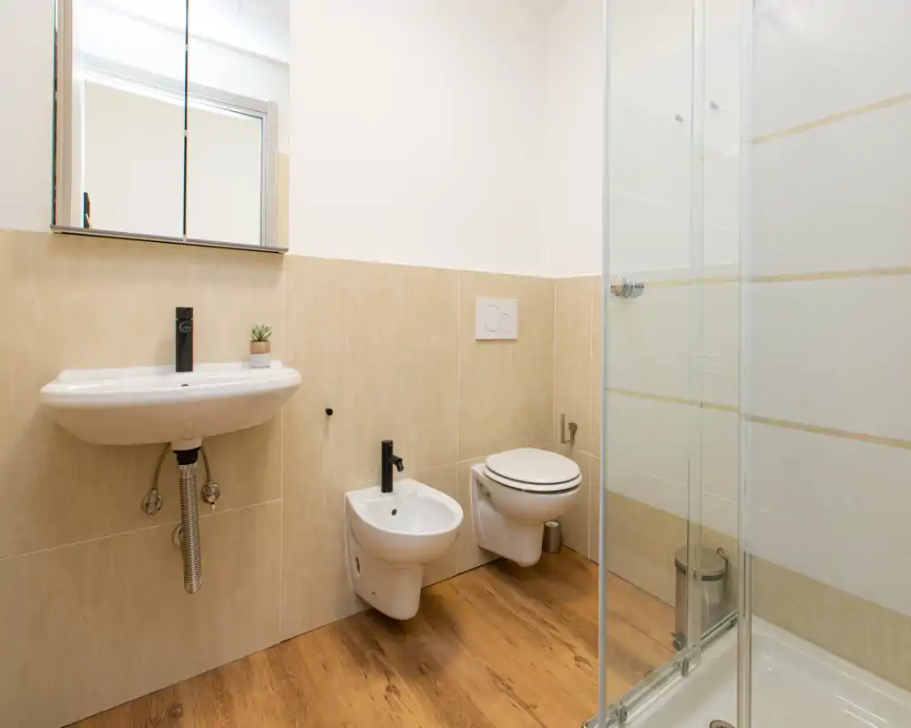 Bagno luminoso e moderno con sanitari sospesi, bidet, lavabo e doccia con box in vetro; pavimento effetto legno e piastrelle neutre per un aspetto pulito e contemporaneo.