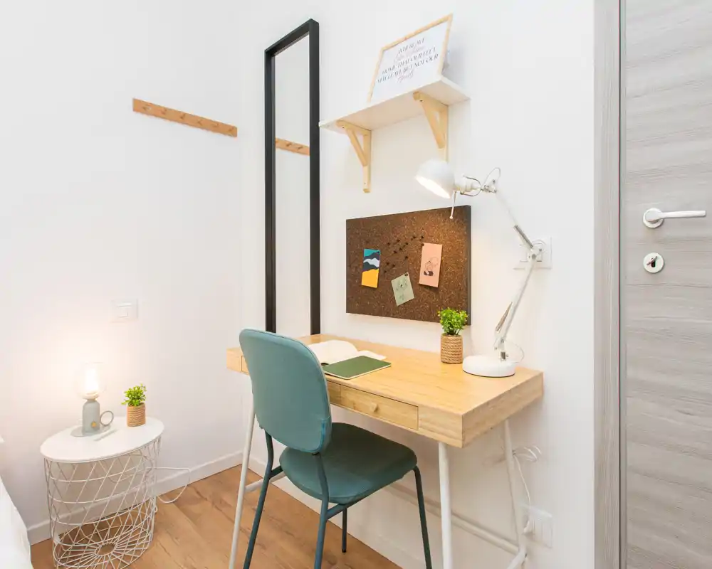 Angolo camera compatto con scrivania in legno e sedia, comodino con lampada e una minima porzione del letto visibile sul bordo dell’immagine. Spazio di lavoro luminoso e ordinato, adatto a brevi soggiorni.
