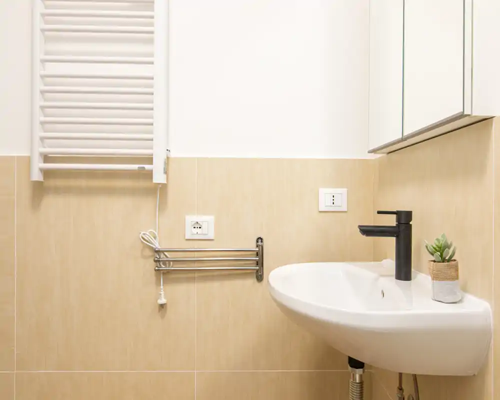 Lavabo da bagno moderno e luminoso con miscelatore nero, porta asciugamani a parete e mobile specchiato. Piastrelle beige neutre e arredo ordinato donano un aspetto pulito e minimale.