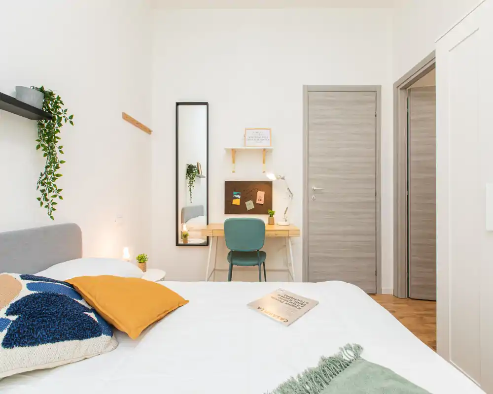 Camera da letto luminosa e moderna con letto ben rifatto, cuscini decorativi, lampada sul comodino e piccolo angolo studio con specchio — ideale per soggiorni confortevoli.
