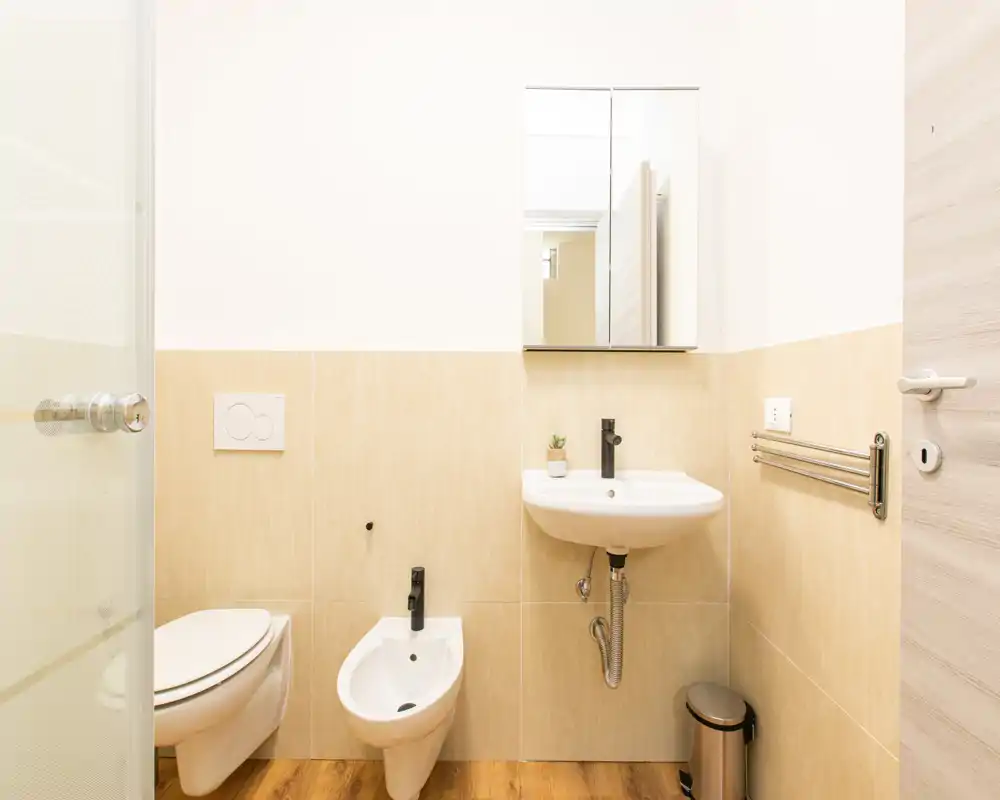 Bagno moderno e luminoso con wc sospeso, bidet e piccolo lavabo sotto un pensile con specchio; piastrelle color caldo e pavimento effetto legno per un aspetto pulito e neutro.