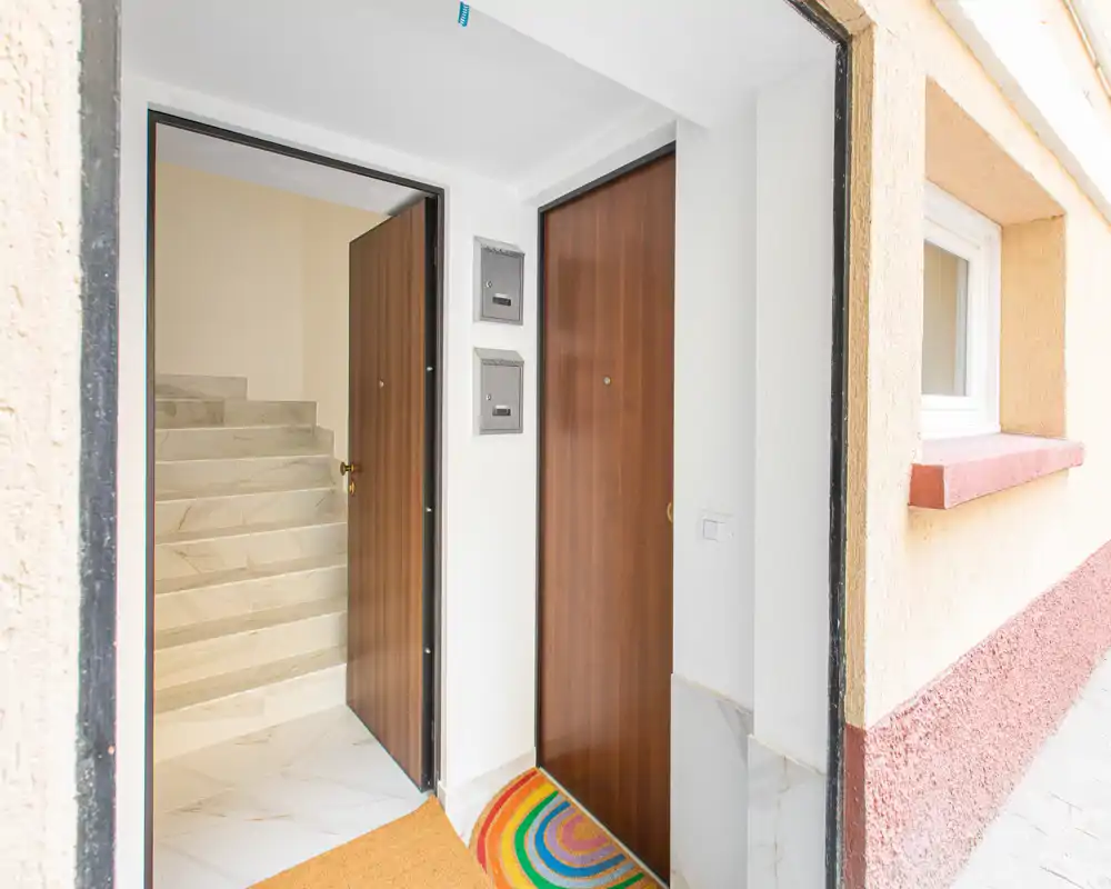 Ingresso luminoso e pulito con due porte in legno, pavimento piastrellato e uno zerbino colorato; si intravede una scala oltre la porta aperta.