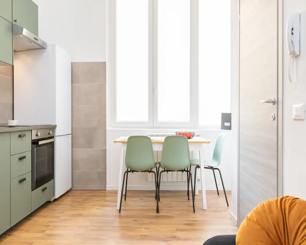 Cucinotto moderno e luminoso con piccolo tavolo da pranzo, sedie verde pastello, pavimento in legno e luce naturale da una grande finestra — perfetto per annunci di monolocali o bilocali.