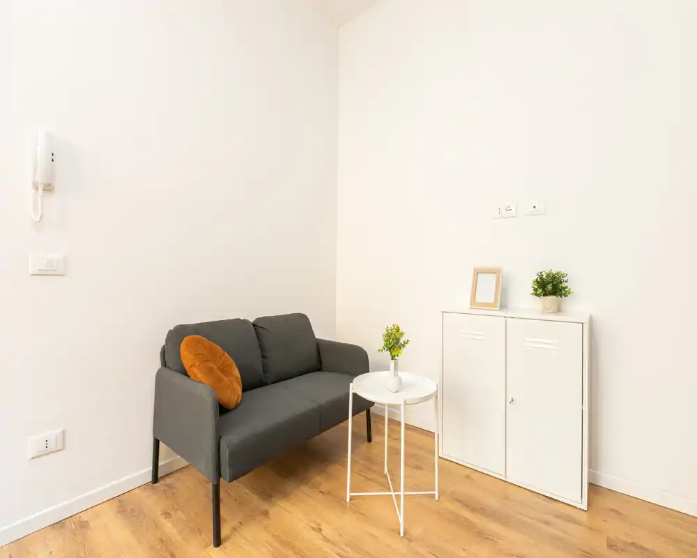 Zona living luminosa e minimal con un divano grigio scuro compatto, tavolino bianco rotondo e un mobiletto semplice — accogliente e ben curata per un piccolo appartamento.
