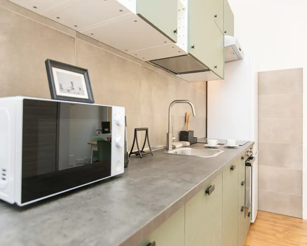 Cucina compatta e moderna con piano di lavoro lungo, lavello integrato, microonde e piano cottura; piastrelle neutre e mobili verdi chiaro creano un ambiente funzionale e rassicurante.