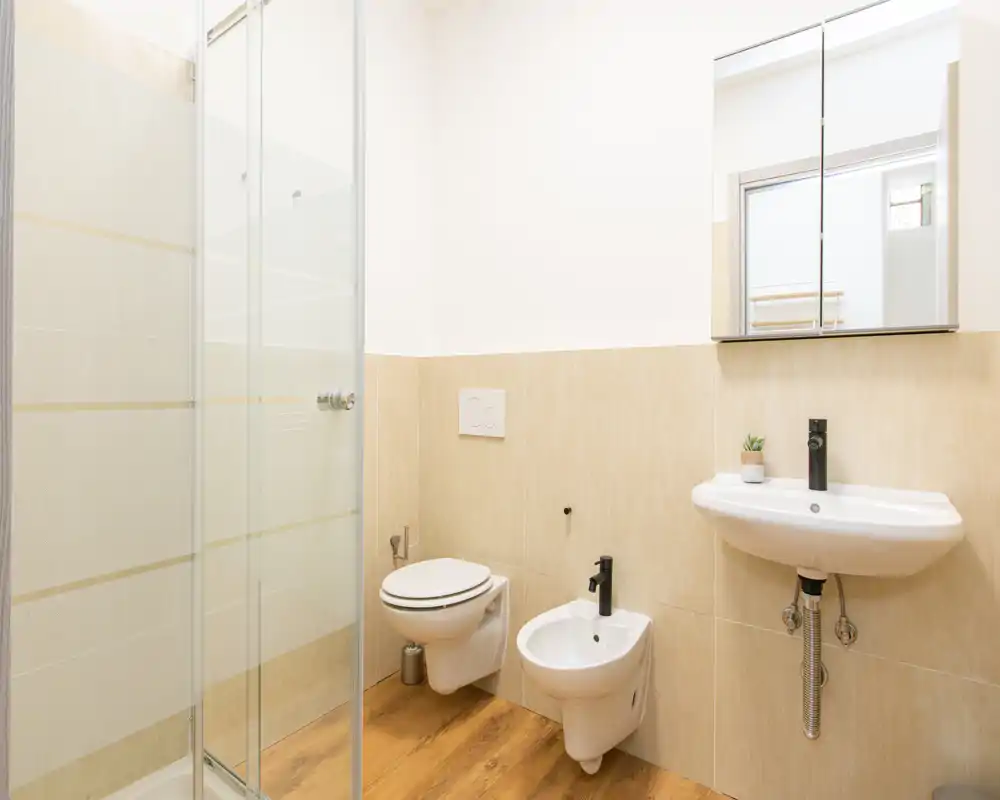 Bagno moderno e luminoso con doccia in vetro, wc sospeso, bidet e lavabo a parete sotto un mobile specchiato.