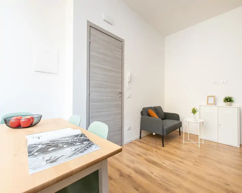Zona giorno luminosa e minimal con poltrona confortevole, tavolini e pavimento in legno chiaro — perfetta per rilassarsi in un appartamento moderno e compatto.