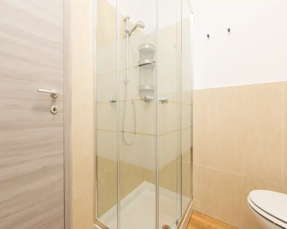 Bagno moderno e compatto con cabina doccia in vetro, soffione e piastrelle beige chiare per un aspetto pulito e minimalista.