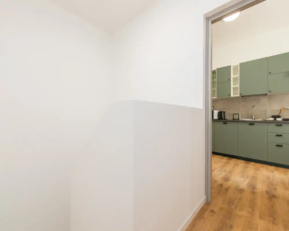 Vista su una cucina compatta con mobili verdi, pavimento effetto legno e paraschizzi neutro, fotografata dall'adiacente scala/ pianerottolo.