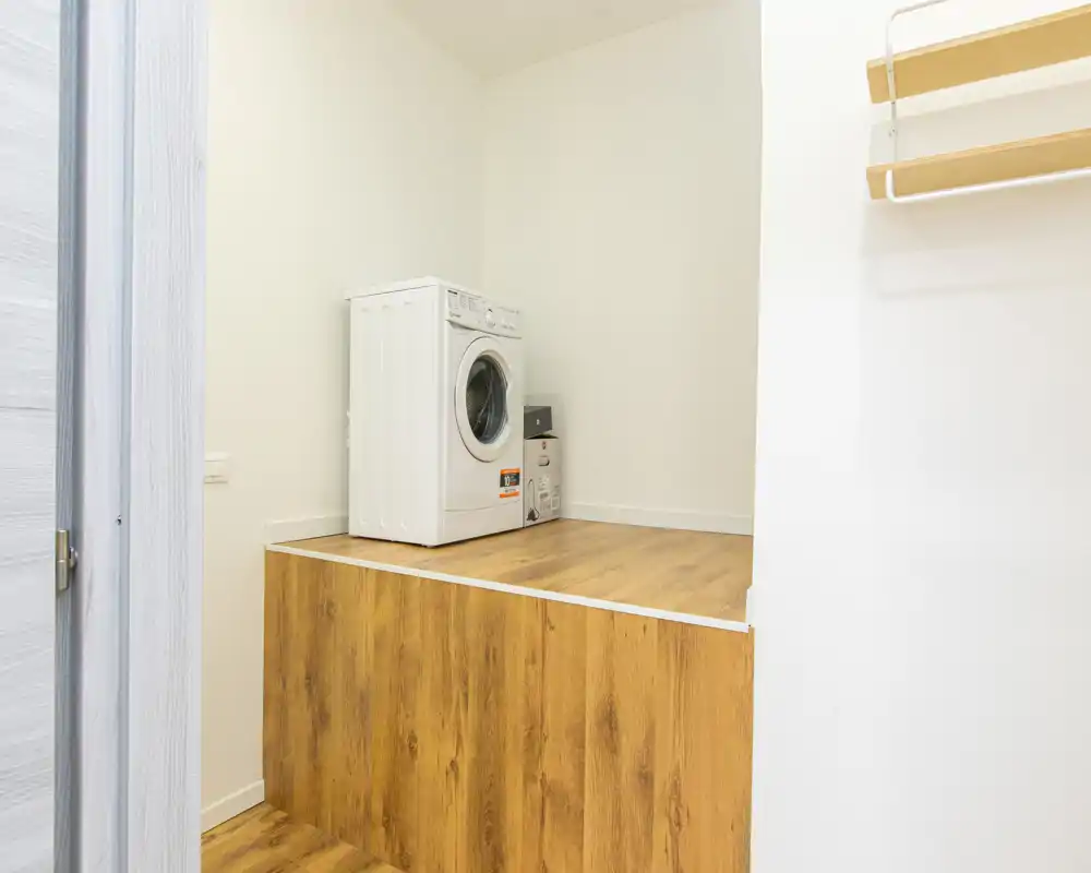 Piccolo angolo lavanderia luminoso con lavatrice a carica frontale su un piano rialzato in legno; spazio pulito, minimale e funzionale ideale per appartamenti compatti.
