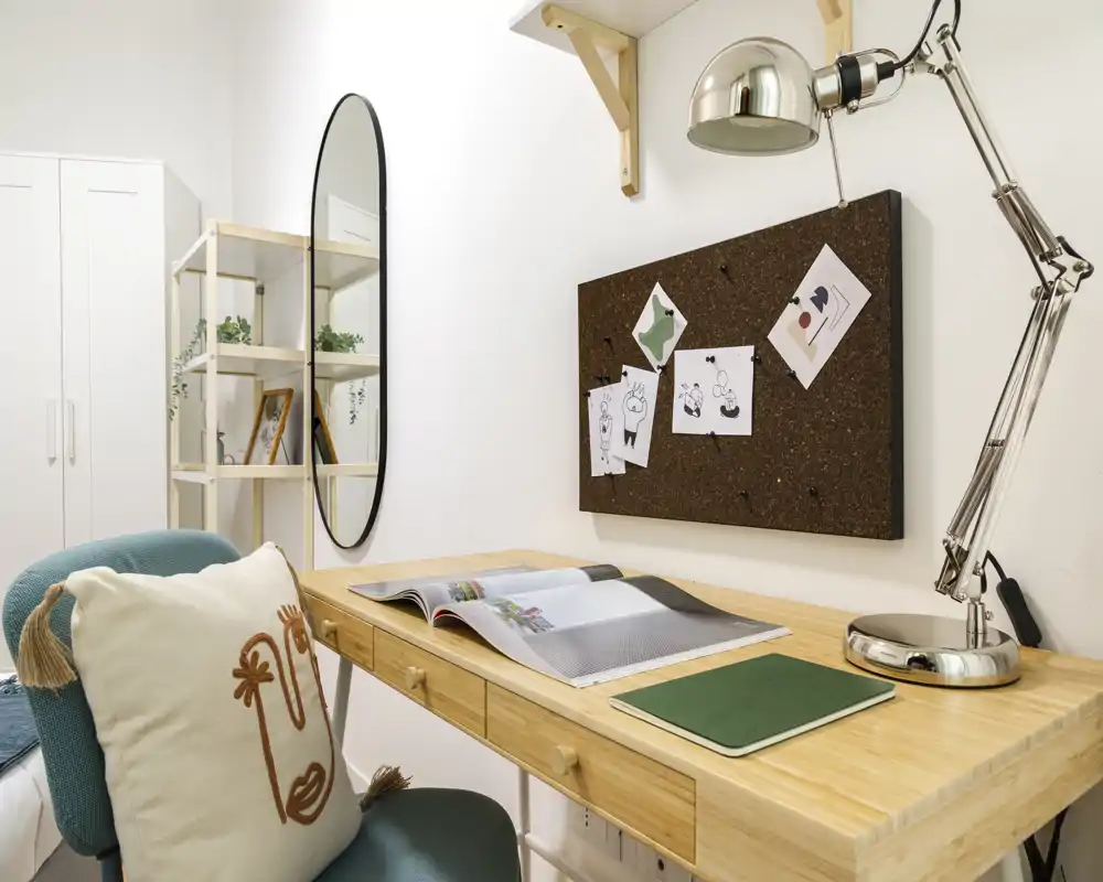 Angolo lavoro ordinato e luminoso con scrivania in legno, lampada regolabile, pannello in sughero per appunti e una sedia comoda con cuscino decorativo.