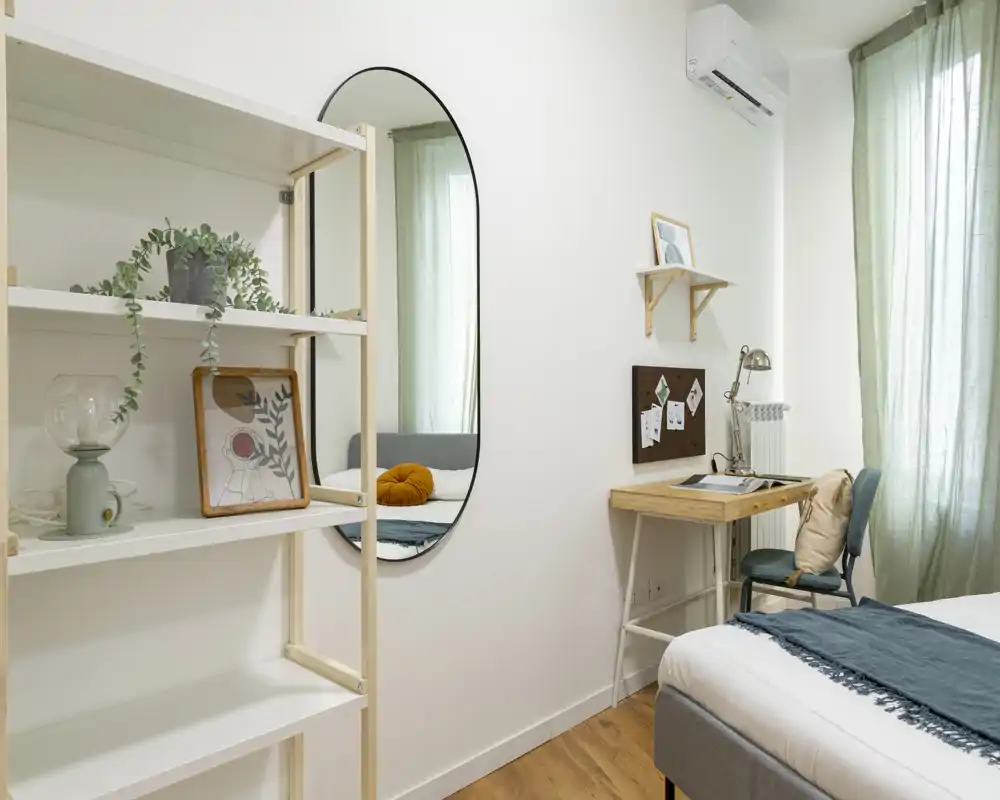 Camera da letto luminosa e moderna con letto rifatto, cuscini decorativi, una scrivania piccola e scaffalatura che creano uno spazio accogliente e funzionale.