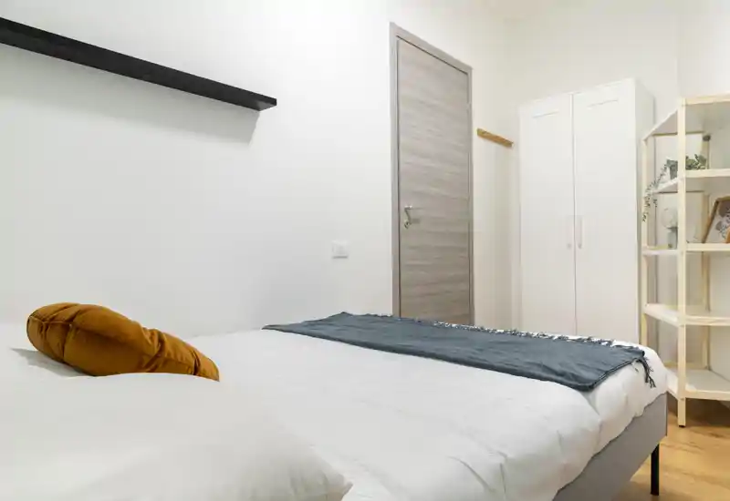 Camera da letto luminosa e minimale con un ampio letto con biancheria bianca, plaid verde acqua e cuscino ocra; armadio e scaffale semplici visibili a destra.