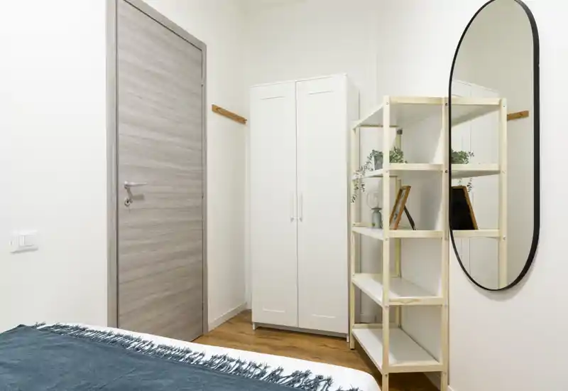 Angolo di camera da letto moderno con bordo del letto visibile, armadio bianco, scaffalatura con piante e grande specchio ovale; luminoso, minimal e ordinato.