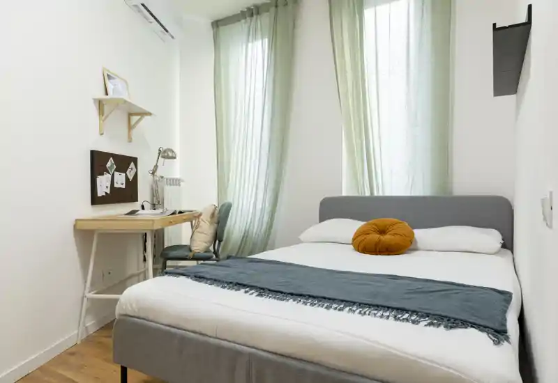 Camera da letto luminosa e ben arredata con un comodo letto matrimoniale, lino chiaro, un cuscino decorativo rotondo e una scrivania vicino alla finestra che lascia entrare luce naturale.