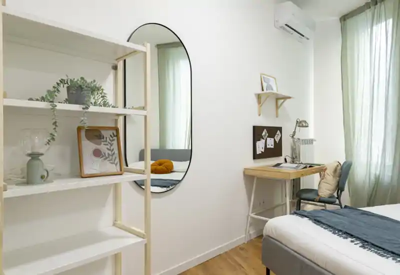 Camera da letto luminosa e moderna con letto rifatto, cuscini decorativi, una scrivania piccola e scaffalatura che creano uno spazio accogliente e funzionale.